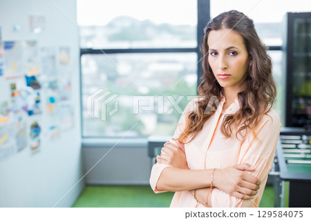 Woman in late twenties crossing arms in office showing pinboard, foosball table, copy space 129584075