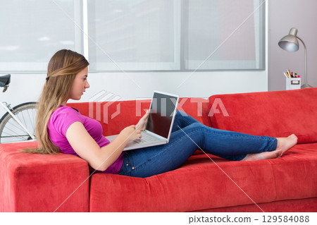 Woman reclining on red sofa in living room typing on silver laptop beside white bicycle 129584088