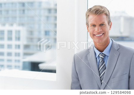 Businessman in suit leaning against window mullion in office overlooking city skyline, copy space 129584098