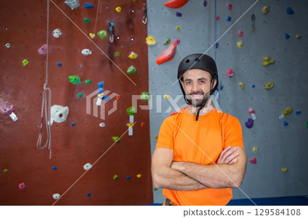 Man standing with arms crossed by climbing rope in climbing gym wearing black helmet, copy space 129584108