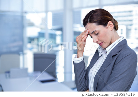 Businesswoman in blazer rubbing forehead at office desk with laptop, papers, pen cup, copy space 129584119