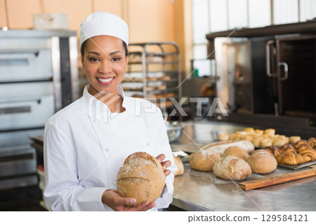 African American woman wearing chef coat and baker cap holding round loaf in bakery kitchen African American woman wearing chef coat and baker cap holding round loaf in bakery kitchen 129584121