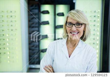 Woman wearing lab coat standing before lit eyeglass display racks in optical shop, copy space 129584123