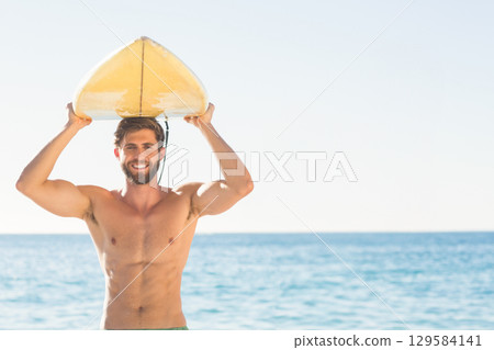 Male surfer standing at water's edge on beach wearing swim trunks holding yellow surfboard overhead 129584141