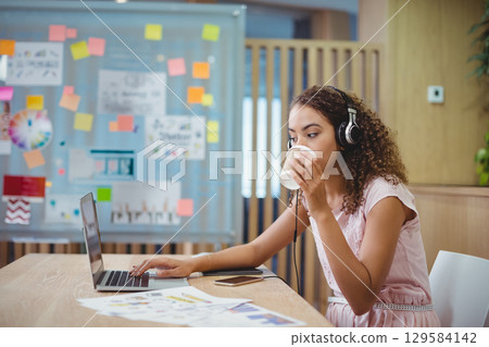 African American woman in pink dress and headphones working on laptop in office, copy space 129584142