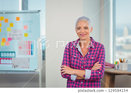 Senior woman standing at office before whiteboard with charts and desk with colored pencils 129584154