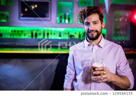 Bearded man holding beer mug at bar counter with neon-lit shelves stocked with bottles, copy space 129584155