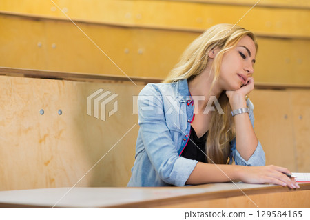 Teenage female student wearing denim shirt sitting writing in notebook at lecture hall, copy space 129584165