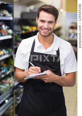 Man in black apron holding pen and spiral-bound notepad while stocking supermarket produce aisle 129584166