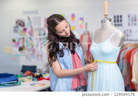 Woman measuring pale blue dress on mannequin in design studio using yellow tape and swatches 129584176