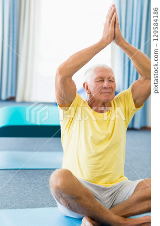 Senior man practicing seated meditation on blue mat in fitness studio with curtains Senior man practicing seated meditation on blue mat in fitness studio with curtains 129584186