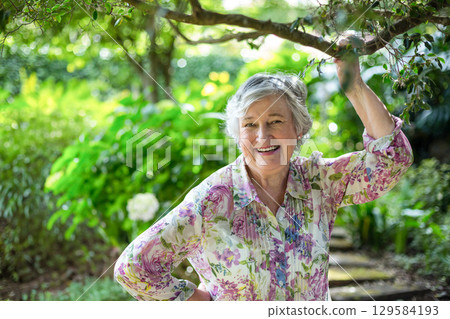 Senior woman standing in garden holding tree branch and wearing floral-print blouse on stone path 129584193