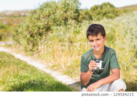 School-aged boy crouching beside dirt path in green shirt and shorts holding binoculars, copy space 129584215