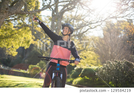 Female cyclist wearing sportswear, helmet and gloves riding bicycle along park path pointing upward Female cyclist wearing sportswear, helmet and gloves riding bicycle along park path pointing upward 129584219