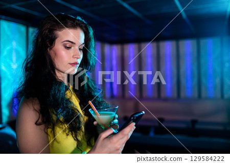 Woman wearing yellow dress holding smartphone and martini glass near bench in lounge, copy space Woman wearing yellow dress holding smartphone and martini glass near bench in lounge, copy space 129584222