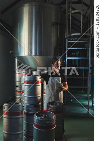 Standing among stainless steel kegs on brewery floor man in his 30s holding tablet Standing among stainless steel kegs on brewery floor man in his 30s holding tablet 129584226