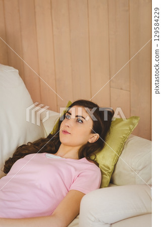 Woman in twenties reclining on white sofa at home with green satin pillow by wood wall 129584229