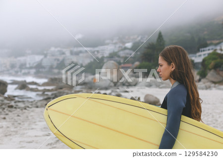 Female surfer standing on foggy beach wearing wetsuit and holding yellow surfboard, copy space Female surfer standing on foggy beach wearing wetsuit and holding yellow surfboard, copy space 129584230