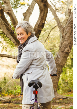 Senior woman standing in wooded park holding bicycle handlebars wearing gray coat and knit scarf Senior woman standing in wooded park holding bicycle handlebars wearing gray coat and knit scarf 129584232