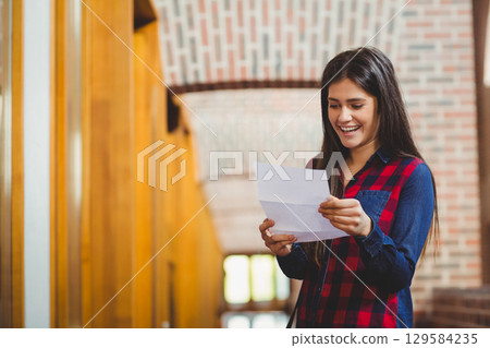 Woman standing in library corridor wearing plaid shirt and reading folded letter, copy space 129584235