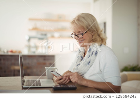 Senior woman sipping coffee while using laptop, smartphone, calculator at kitchen table, copy space 129584242