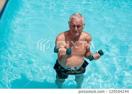 Senior man exercising with dumbbells near pool deck edge in bright sun wearing floral swim trunks Senior man exercising with dumbbells near pool deck edge in bright sun wearing floral swim trunks 129584244