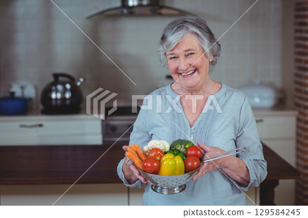 Senior woman smiling while holding metal colander of fresh vegetables in home kitchen 129584245