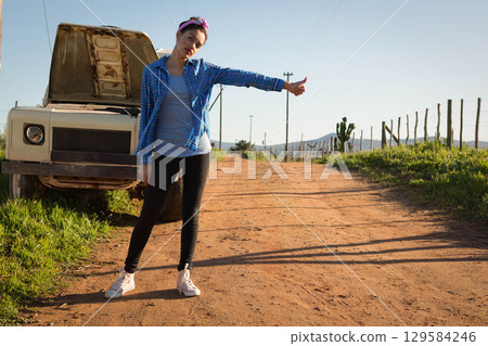 Woman hitchhiking beside white pickup truck with open hood on rural dirt road, copy space Woman hitchhiking beside white pickup truck with open hood on rural dirt road, copy space 129584246