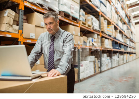 Man in his 50s wearing tie typing on laptop at box in warehouse aisle, copy space 129584250