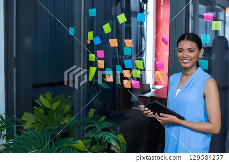 Asian woman holding digital tablet by glass partition with sticky notes in office, copy space 129584257