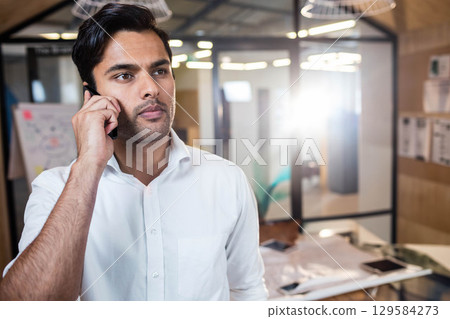 Asian man standing in modern office holding smartphone to ear beside flip chart 129584273