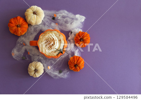 Halloween themed flat lay with a pumpkin latte, decorative pumpkins and spiderwebs on a purple background. Halloween themed flat lay with a pumpkin latte, decorative pumpkins and spiderwebs on a purple background. 129584496