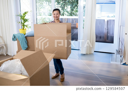 Mid adult Asian woman carrying cardboard box inside bright living room with bubble wrap 129584507