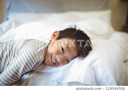 Asian boy lying in bedroom wearing striped pajama top, smiling and resting head on white pillow 129584508