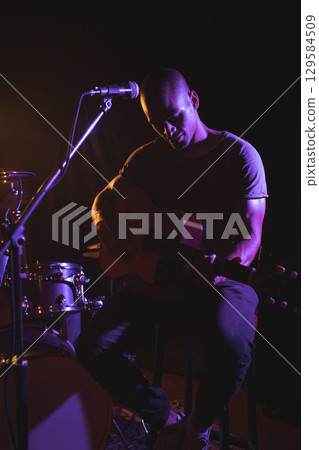 African American male musician playing acoustic guitar on stage under purple lights with mic stand 129584509
