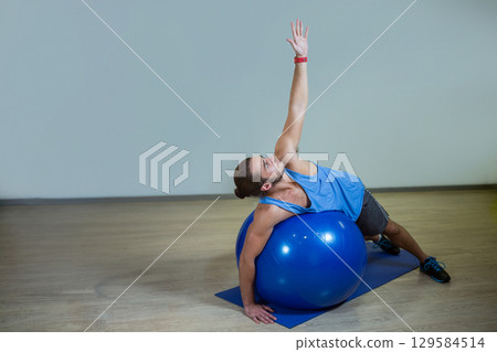 Man wearing blue top and shorts stretching sideways over blue stability ball on mat in gym 129584514