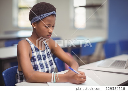African American girl student writing in open notebook at classroom table with silver laptop nearby African American girl student writing in open notebook at classroom table with silver laptop nearby 129584516