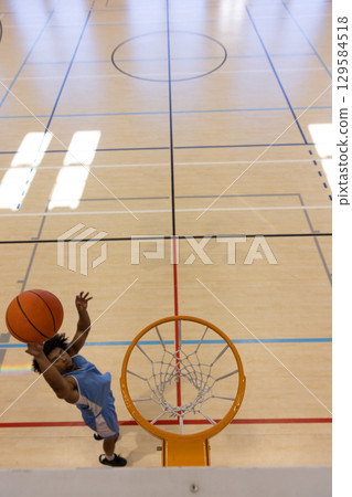 African American man in blue jersey leaping and dunking orange basketball into hoop on gym floor 129584518
