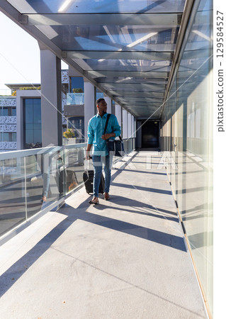 African American male checking wristwatch walking glass walkway pulling suitcase and navy duffel 129584527