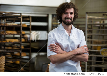 Mid adult man baker crossing arms and smiling in bakery area with metal racks of bread Mid adult man baker crossing arms and smiling in bakery area with metal racks of bread 129584532