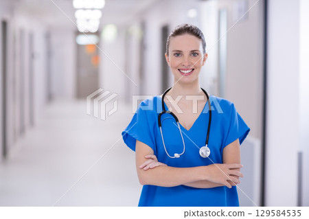 Female healthcare worker standing in hospital corridor wearing blue scrubs and holding stethoscope 129584535