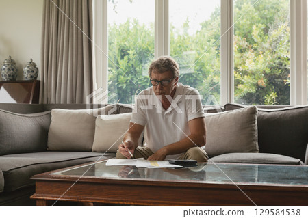 Senior man wearing polo shirt sitting in living room using highlighter on papers near smartphone Senior man wearing polo shirt sitting in living room using highlighter on papers near smartphone 129584538