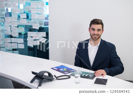 Male businessman reviewing printed charts at reception desk with tablet and phone in office Male businessman reviewing printed charts at reception desk with tablet and phone in office 129584541