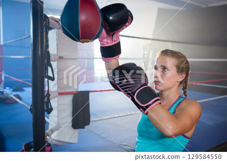 Female athlete striking speed bag in ring corner in teal top and black-and-pink gloves, copy space 129584550