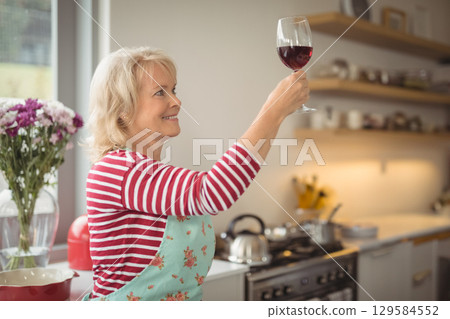Senior woman wearing apron raising wine glass at kitchen window with vase of flowers and utensils Senior woman wearing apron raising wine glass at kitchen window with vase of flowers and utensils 129584552