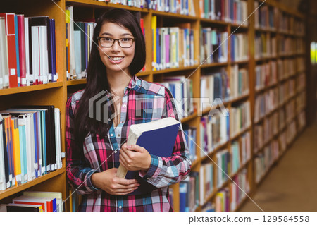 Asian woman in plaid shirt leaning against bookshelf holding blue book in library aisle, copy space 129584558