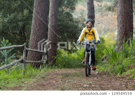 Male cyclist wearing yellow helmet backpack mountain biking along forest trail by wooden fence 129584559