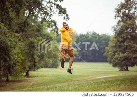 Man jumping wearing bright yellow hoodie and brown shorts twisting in grassy park along dirt path 129584566