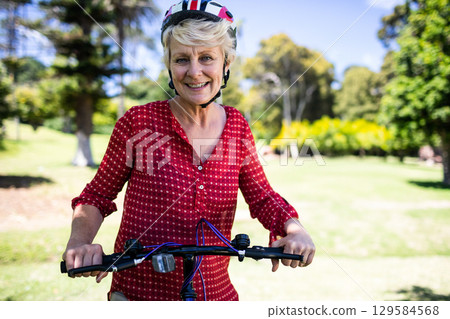 Senior woman holding bicycle handlebars in grassy park wearing red blouse, bicycle helmet Senior woman holding bicycle handlebars in grassy park wearing red blouse, bicycle helmet 129584568