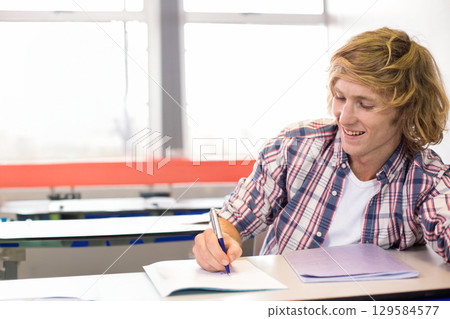Male student writing in notebook with blue pen, closed binder on lecture hall desk, copy space Male student writing in notebook with blue pen, closed binder on lecture hall desk, copy space 129584577